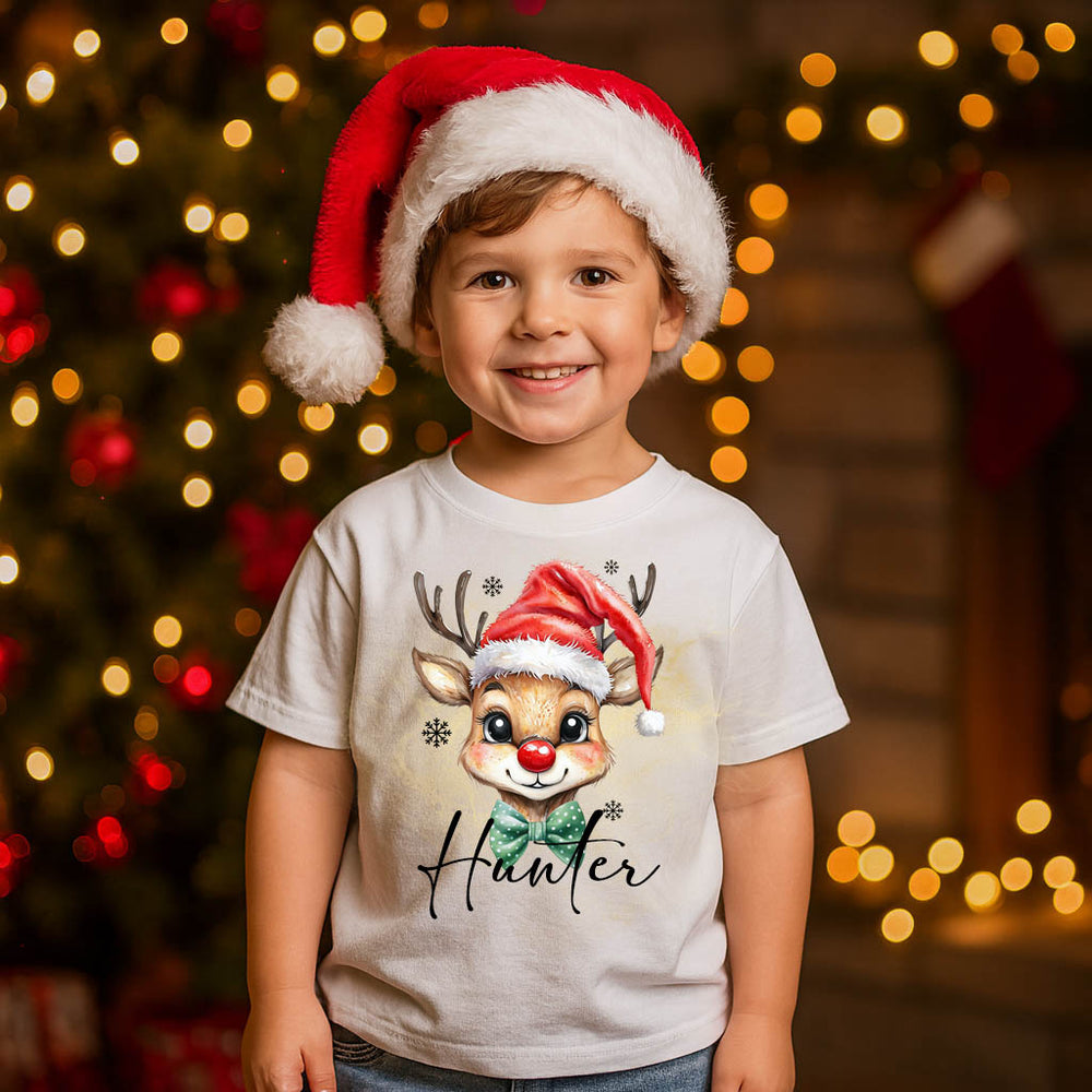 Child wearing a Santa hat and a t-shirt with a reindeer design in front of a Christmas tree.