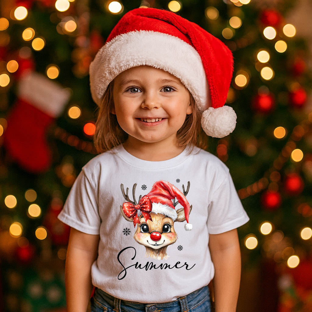 Child wearing a Santa hat and a shirt with a reindeer design in front of a Christmas tree.