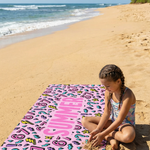 Child sitting on a colorful towel at the beach with ocean and palm trees in the background